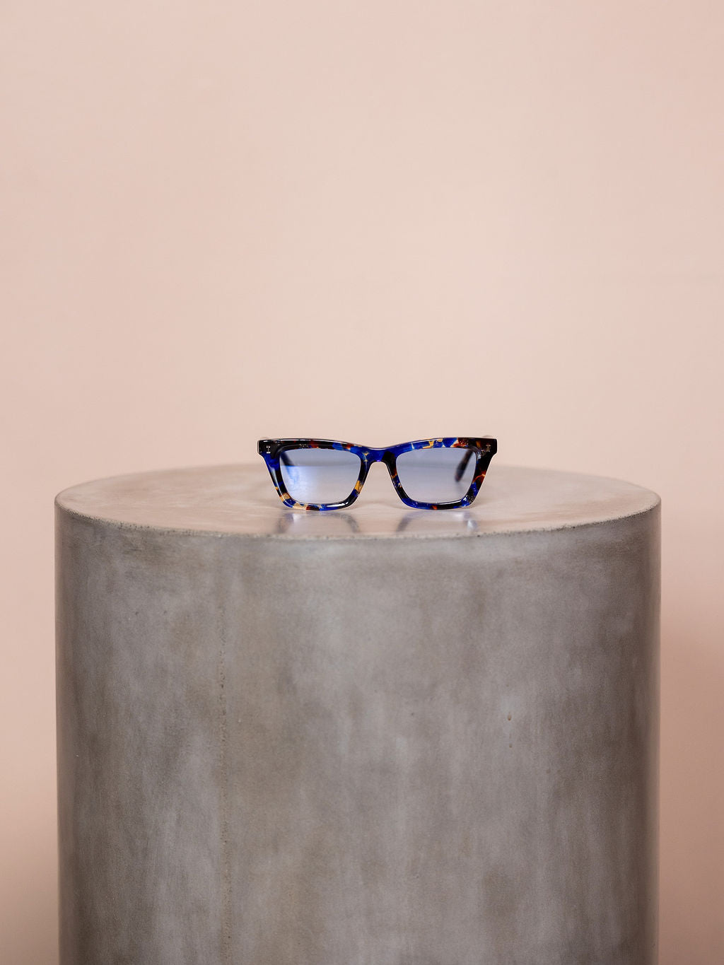 Patterned blue sunglasses on podium against pink background