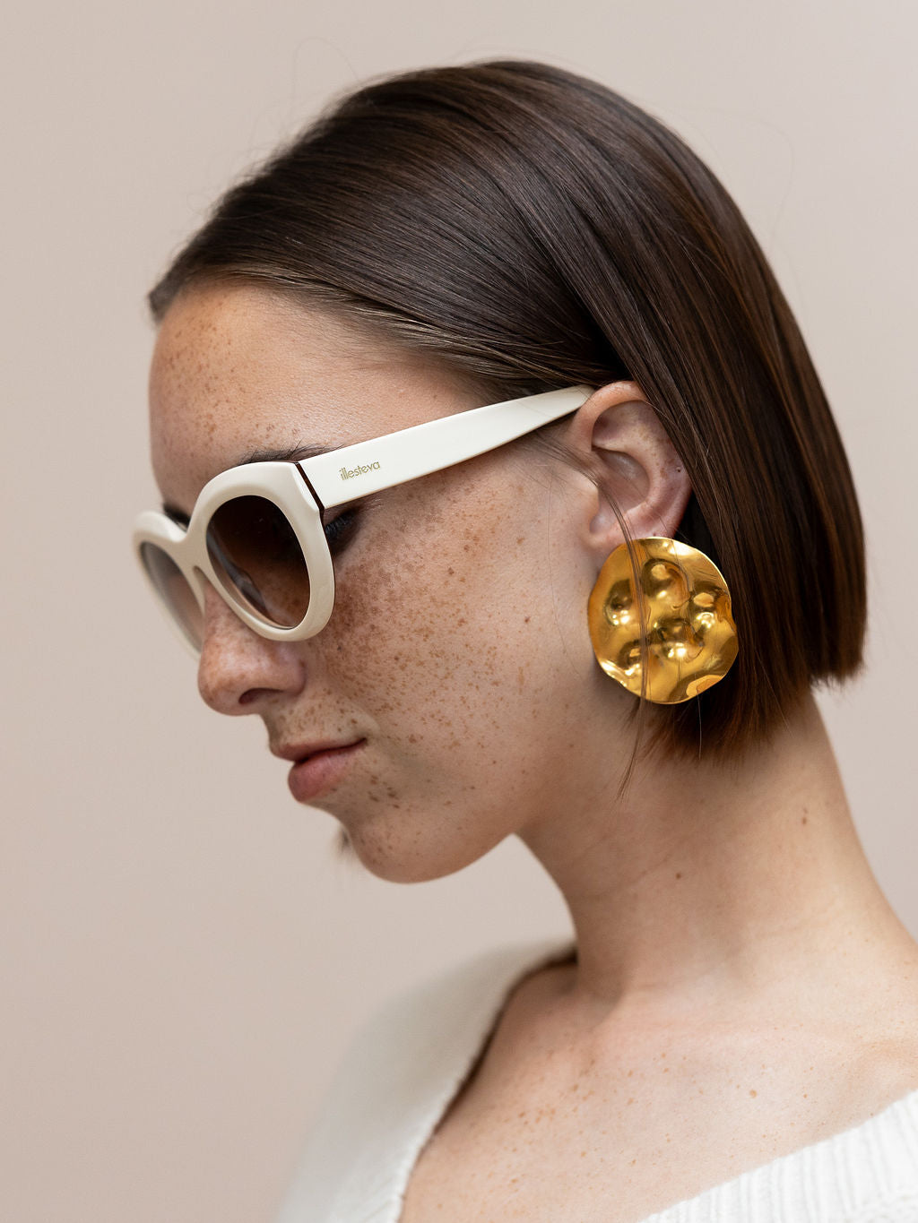 Woman wearing cream sunglasses and textured gold disc earrings against pink background
