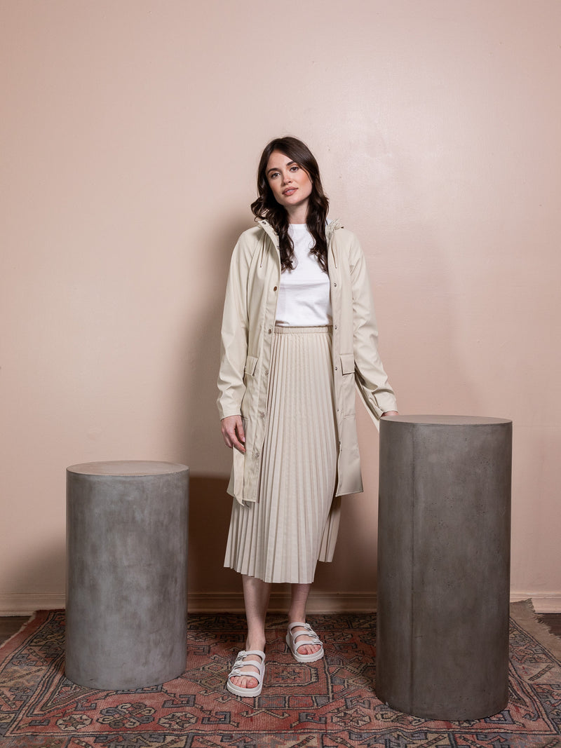 Woman in tan rain jacket and tan skirt against pink background