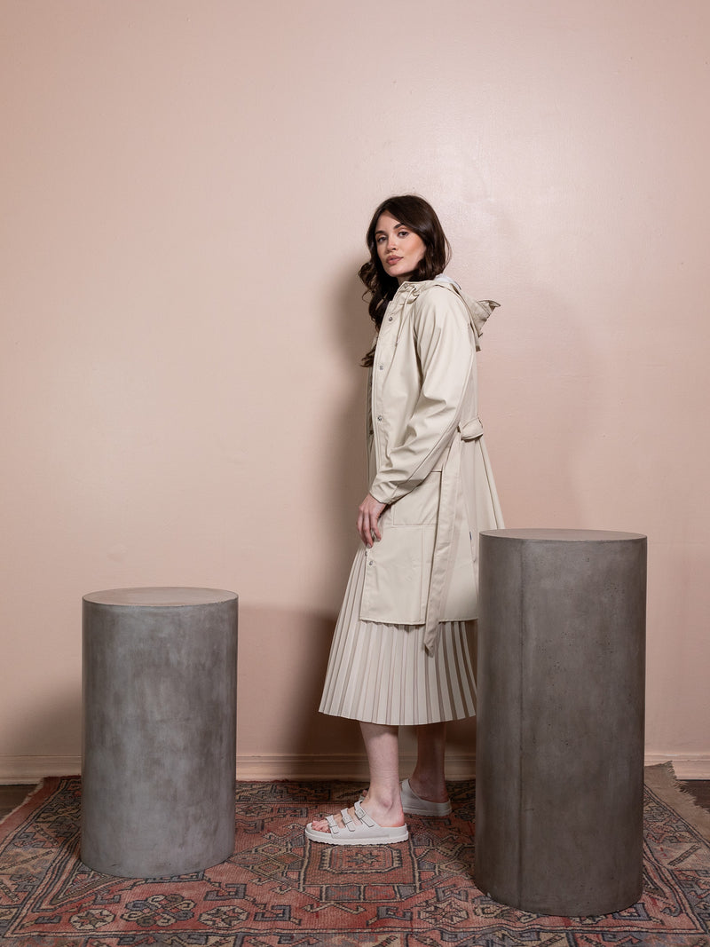 Woman in tan rain jacket and tan skirt against pink background