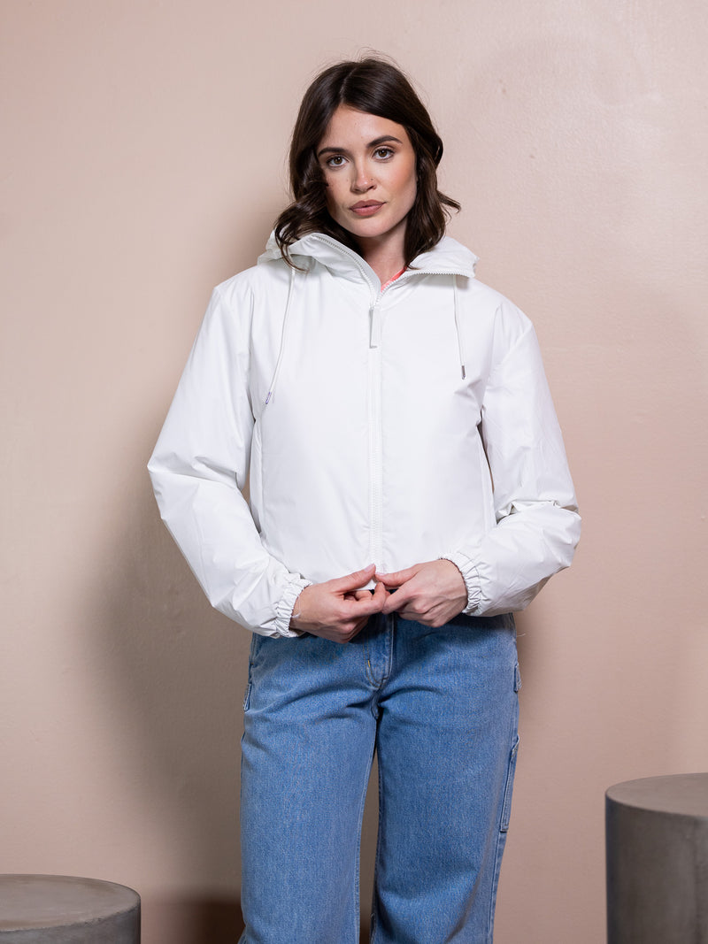 Woman in pink top, white rain jacket, and blue jeans against pink background