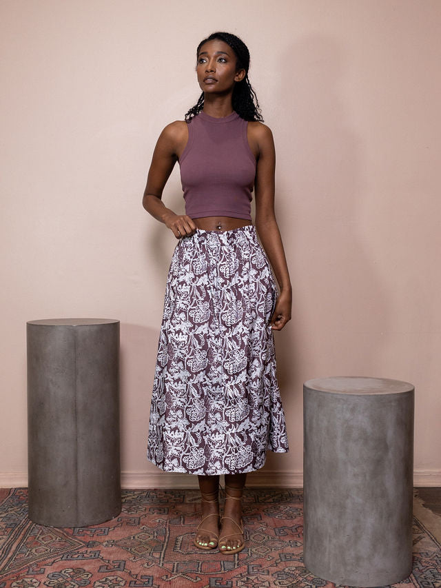 Woman wearing maroon cropped tank top and maroon and white patterned skirt against pink background