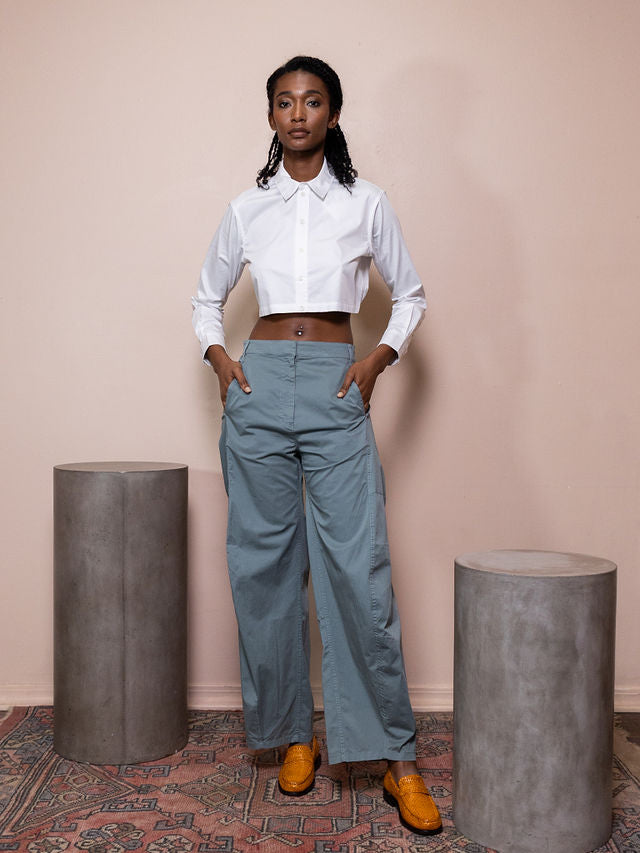 Woman wearing white cropped top and teal pants against pink background