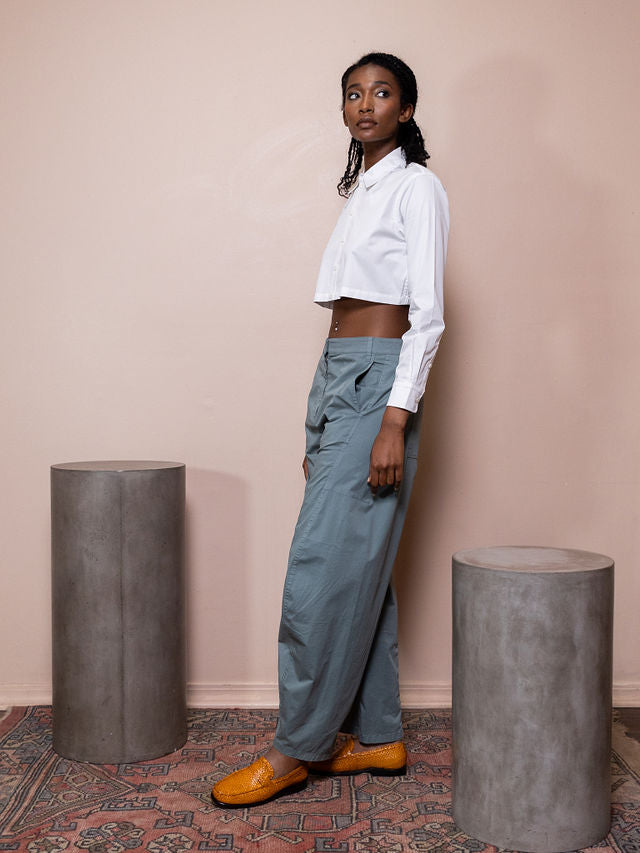 Woman wearing white cropped top and teal pants against pink background