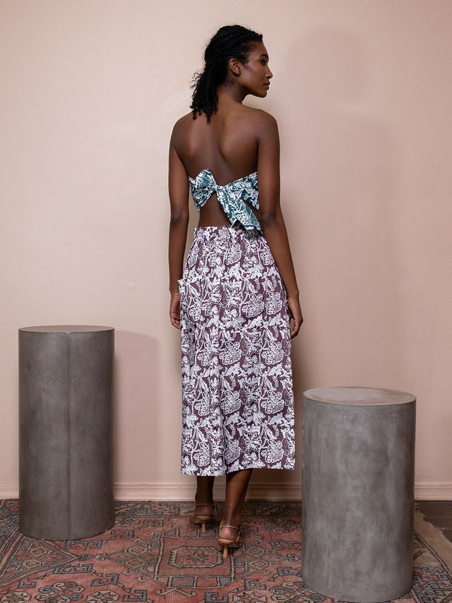 Woman wearing teal and white patterned bandeau top and maroon and white patterned skirt against pink background