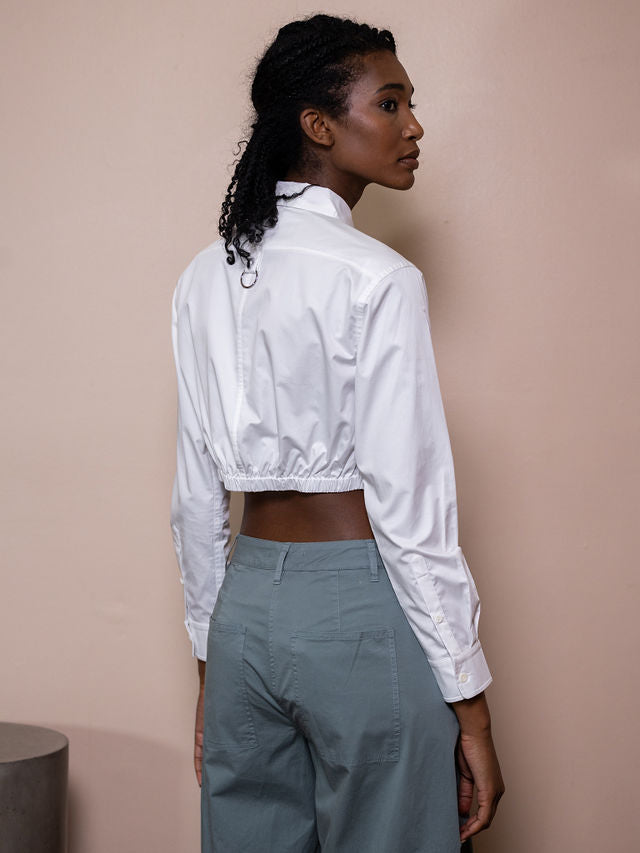 Woman wearing white cropped top and teal pants against pink background